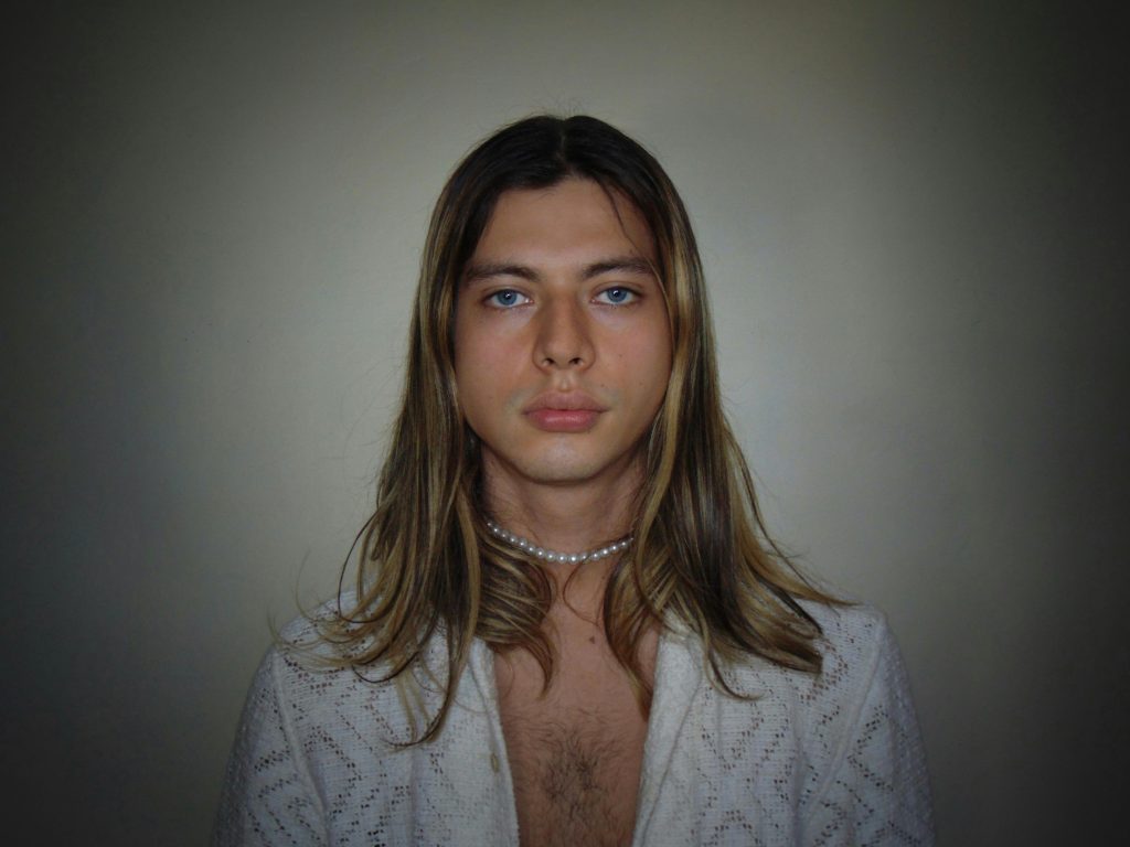 Close-up portrait of a person with long hair and a pearl necklace against a neutral background.